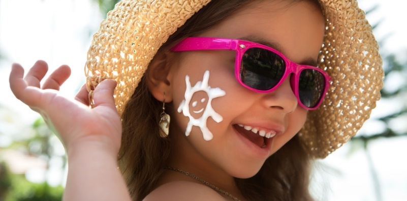 A young girl wearing a hat and sunglasses with a sun painted on her face in sun cream.