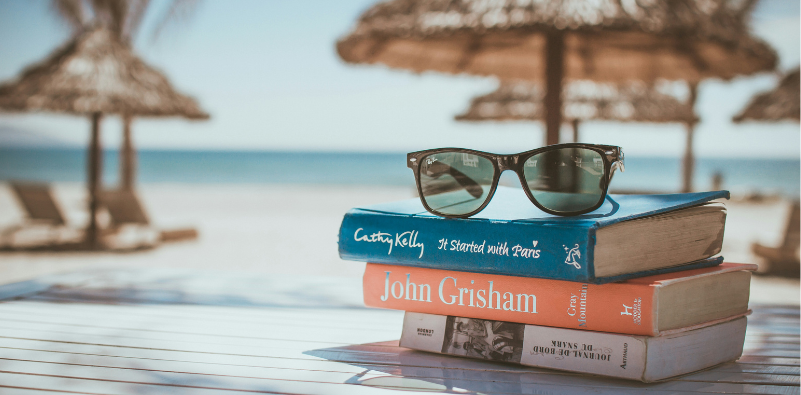 A pair of Ray-Ban sunglasses resting on a pile of books by the beach.
